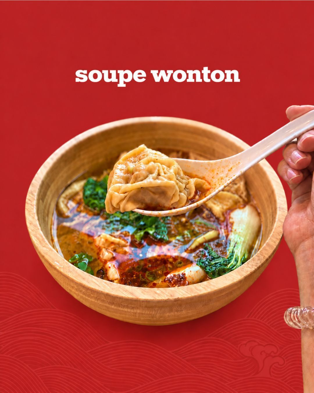 Soupe wonton servie dans un bol en bois avec un ravioli tenu dans une cuillère, sur fond rouge.